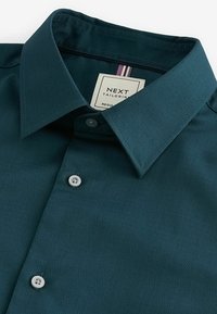 Sötét teal színű, gombos ing, mely textúrált anyagból készült, klasszikus gallérral rendelkezik, és rózsaszín részletekkel ellátott márkalábjegyzékkel rendelkezik. Két fehér gomb látható.