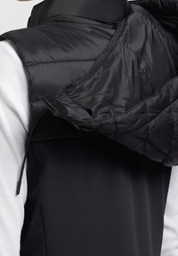 Schwarze Steppweste mit Reißverschluss, gestepptem Innenfutter und weichem Futter, über einem weißen, gerippten Langarmshirt getragen.