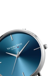Orologio rotondo in argento con un quadrante blu sfumato, marker delle ore e lancette argentati. Nome del brand "Victoria Hyde" mostrato in bianco.