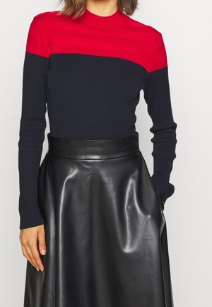 Femme portant un pull rayé rouge et noir à manches longues, rentré dans une jupe en cuir noire taille haute, mains le long du corps.