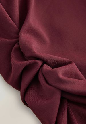 Bordová látka s hladkým texturem, s drapérie a detailem ve tvaru twist, která vyjadřuje hloubku a bohatství barvy.