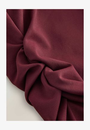 Bordová látka s hladkým texturem, s drapérie a detailem ve tvaru twist, která vyjadřuje hloubku a bohatství barvy.