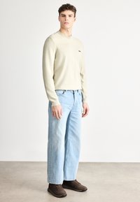 Bézs színű, hosszú ujjú, bordázott textúrájú ing kis logóval, világoskék, laza szabású farmerrel és barna tornacipővel párosítva.