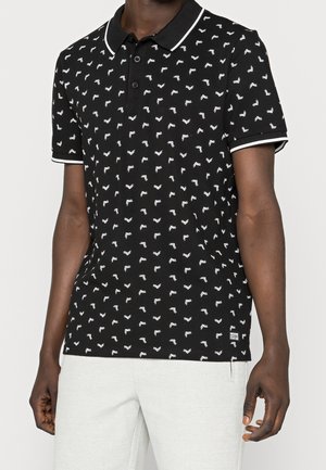 Uomo che indossa una polo nera a maniche corte con piccoli motivi a freccia bianchi e bordo bianco, abbinata a pantaloni grigio chiaro.