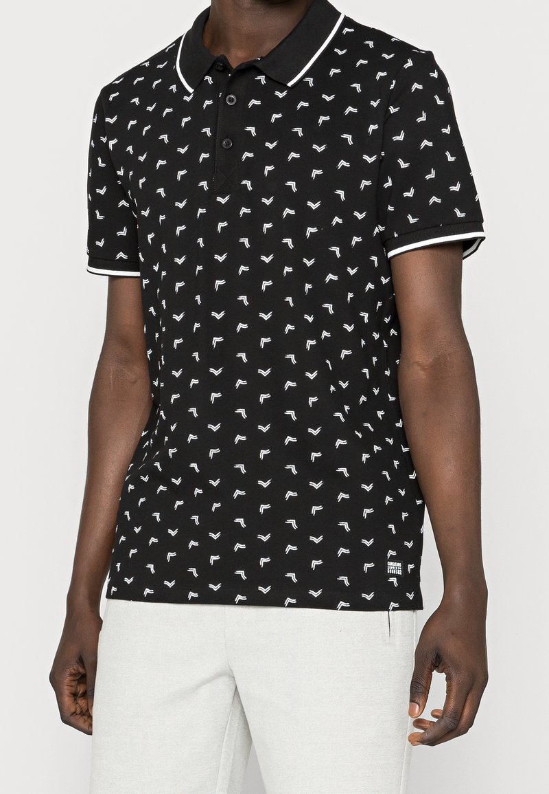 Homme portant un polo noir à manches courtes avec de petits motifs de flèches blanches et une bordure blanche, associé à un pantalon gris clair.