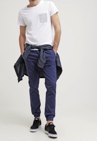 Man in een wit T-shirt met een grijze zak, marineblauwe joggerbroeken, zwarte sneakers en een gestreepte marineblauwe shirt om zijn taille geknoopt.