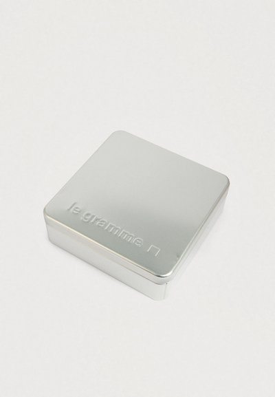 Mali srebrni metalni limeni kutijica s zaobljenim rubovima, utisnuta s tekstom "le gramme" i kvadratnim logotipom na vrhu, na običnoj bijeloj pozadini.