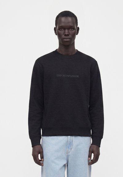 Homme portant un sweat-shirt noir Emporio Armani et un jean bleu clair, se tenant devant un fond clair uni.