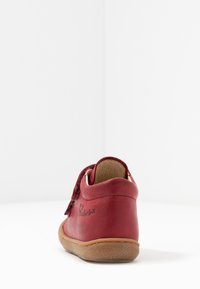 Chaussure en cuir rouge avec une sangle en daim, design incurvé et semelle en caoutchouc texturé. Présente des accents cousus et un logo sur le talon.