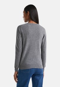 Kobieta z długimi ciemnymi włosami w prostym szarym swetrze i niebieskich dżinsach, stojąca tyłem na białym tle.