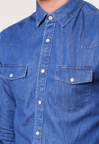 Sininen farkkupaita, jossa on kärkipartainen kaulus, kaksi rintataskua, napit edessä ja tekstuuri-pinta.