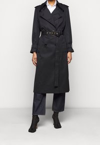 Czarny płaszcz trench z podwójnymi guzikami, paskiem w talii i naramiennikami, w połączeniu z dżinsowymi spodniami typu wide-leg i czarnymi botkami na obcasie.