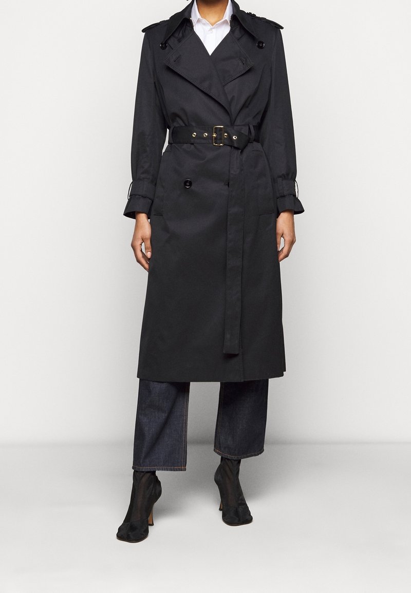 Czarny płaszcz trench z podwójnymi guzikami, paskiem w talii i naramiennikami, w połączeniu z dżinsowymi spodniami typu wide-leg i czarnymi botkami na obcasie.
