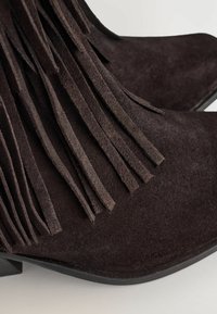 Botines de ante marrón con detalles de flecos largos, punta cuadrada y suela plana. La textura parece suave y aterciopelada.