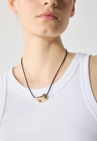 Pandantiv din metal de culoare aurie pe un colier din sfoară neagră, având o formă organică cu o suprafață netedă și reflectantă, purtat deasupra unui top alb cu dungi.