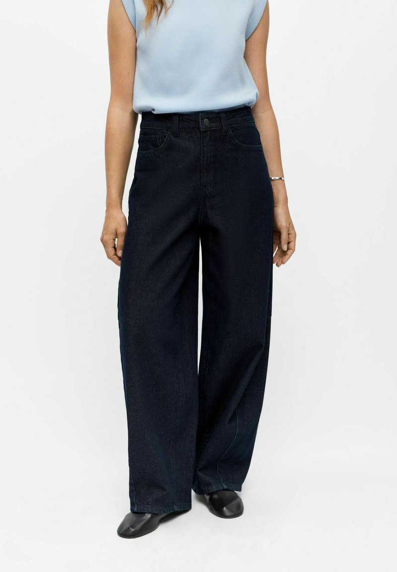 Femme portant un haut sans manches bleu clair, un jean large taille haute de couleur foncée, des chaussures noires et un bracelet fin au poignet gauche.