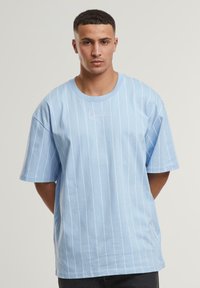 Light blue short-sleeve t-shirt with white vertical stripes and a round neckline. Features a simple white signature graphic on the chest.