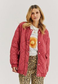 Prešívaná bordová bunda so zapínaním na cvoky a bočnými vreckami, nosená cez biele tričko s grafikou a nohavice s leopardím vzorom. Mäkká textúra.