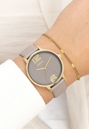 Montre analogique grise avec des accents dorés. Grands marqueurs numériques à 12 et 6 heures. Bracelet en cuir lisse gris. Délicate bracelet en chaîne dorée au poignet.