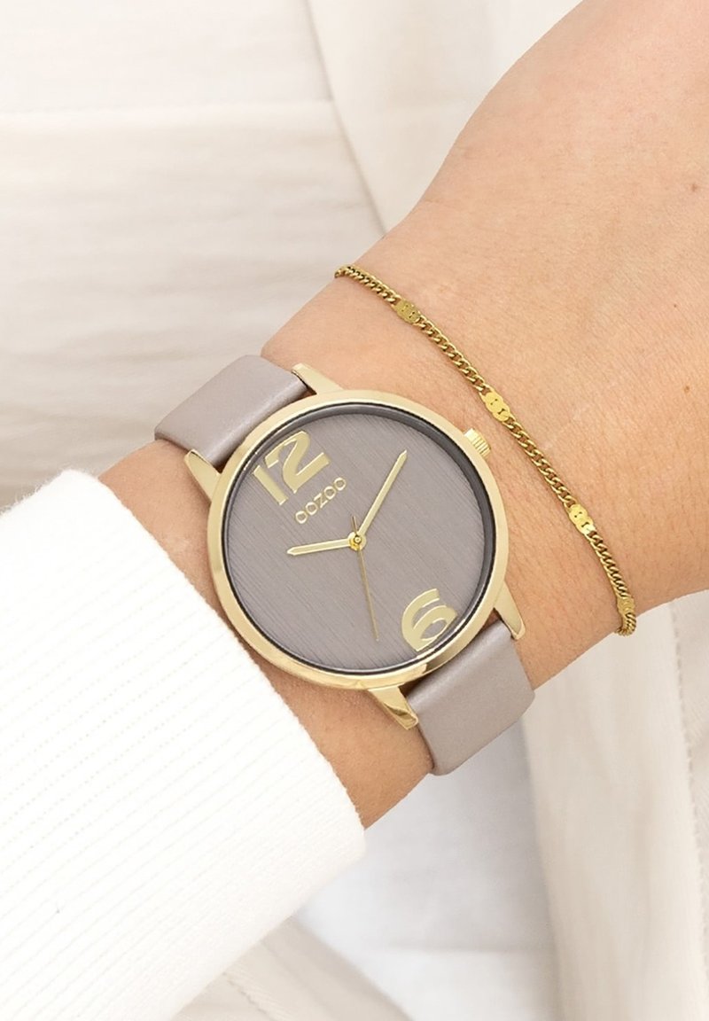 Montre analogique grise avec des accents dorés. Grands marqueurs numériques à 12 et 6 heures. Bracelet en cuir lisse gris. Délicate bracelet en chaîne dorée au poignet.