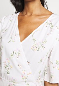 Robe portefeuille blanche ornée d'un motif floral avec des fleurs roses et jaunes et des feuilles vertes. Manches courtes et décolleté en V profond.