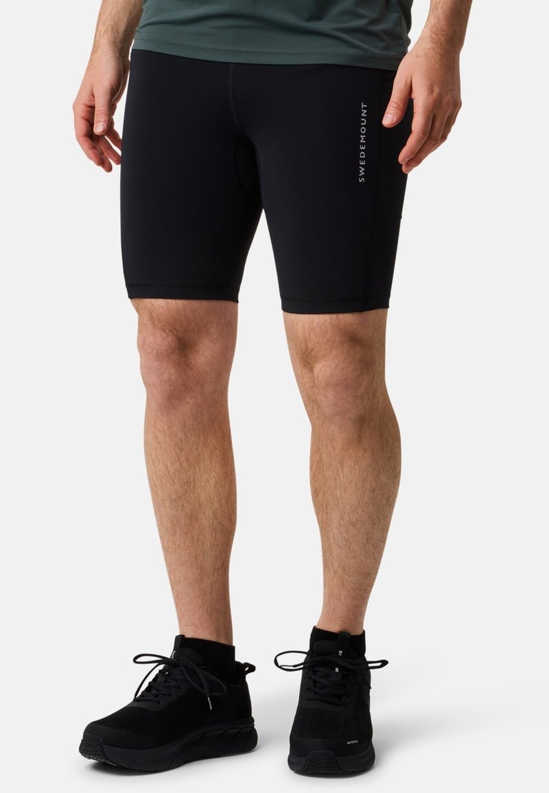 Svarta tajta sportshorts i伸伸ja material, med en sidologotyp och mjuk yta, ihop med svarta joggingskor.