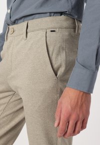 Pantalones beige texturizados con bolsillos laterales, con cierre de botón y pequeños acentos negros, combinados con una camisa de cuello azul claro.