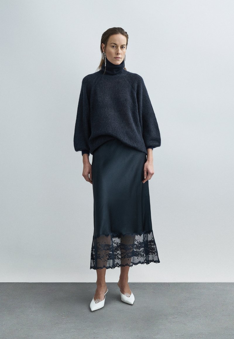 Dark knit sweater with a high collar, paired with a satin midi skirt featuring a lace trim at the hem. White pointed mules complete the look.