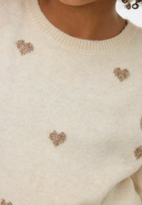 Slonovinově bílý pletený svetr z měkkého materiálu, zdobený roztroušenými hnědými vzory ve tvaru srdcí a kulatým žebrovaným výstřihem.