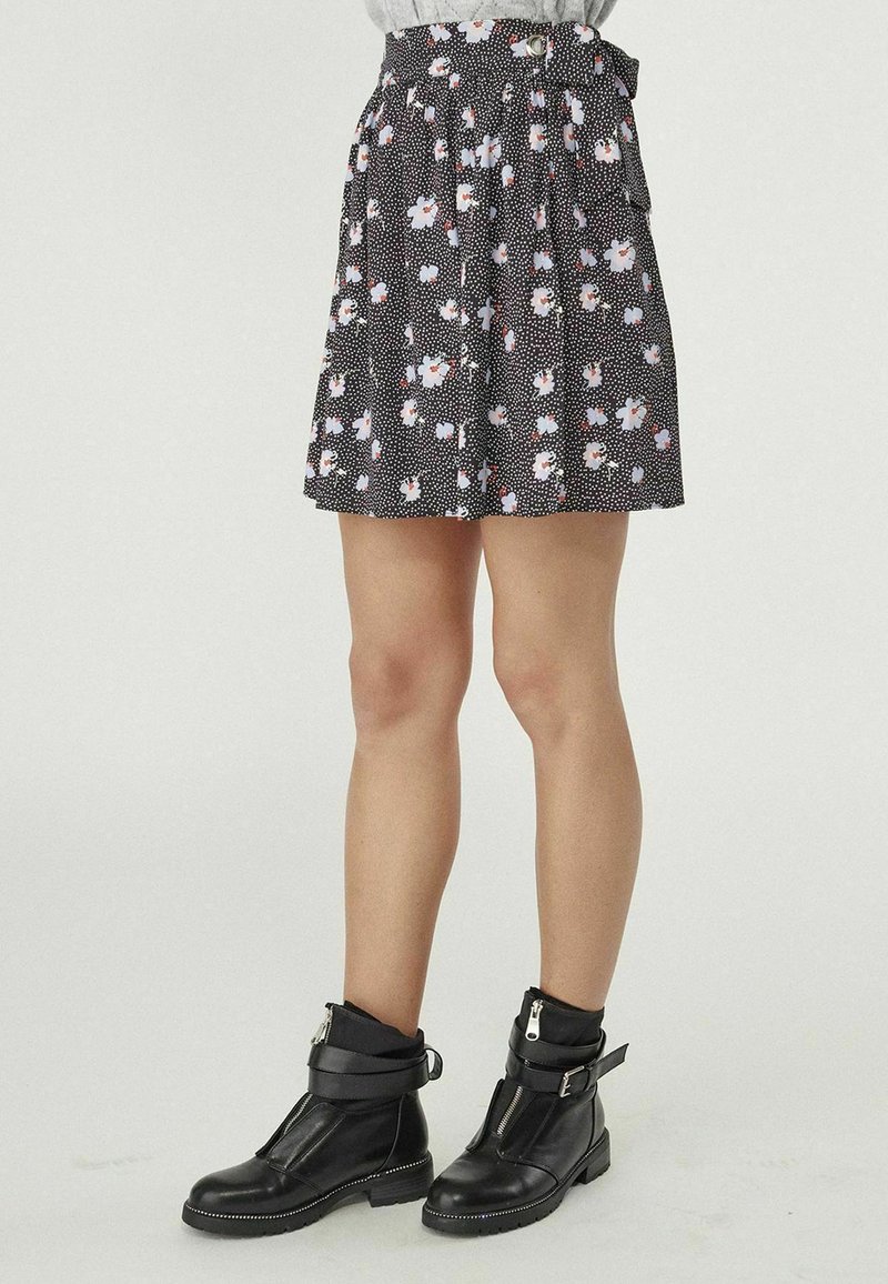 Femme portant une jupe noire fleurie avec de petites fleurs blanches et roses, associée à des bottines noires avec fermetures éclair et boucles.