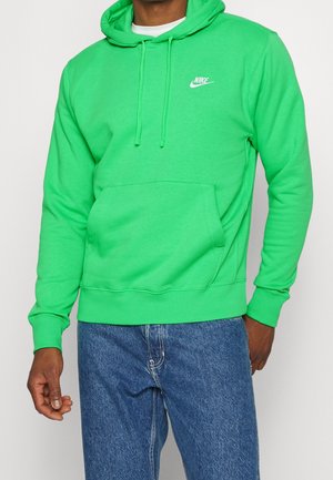 Mężczyzna w jaskrawozielonym bluzie Nike z kapturem i kieszenią z przodu oraz w niebieskich dżinsach, stojący na tle białego, jednolitego tła.