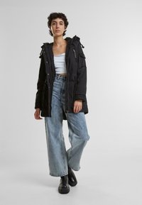 Svart parka med hette, store lommer og glidelåsdetaljer, kombinert med lyseblå vide bukser og svarte ankelstøvler.