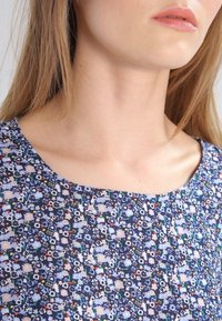 Close-up of a woman’s neck and shoulders wearing a blue floral-patterned top with small multicolored flowers.