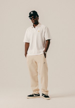 Vīrietis, kurš valkā melnu beisbola cepuri, melnus saulesbrilles, baltu polo kreklu, bēšas sporta bikses un melnas sporta kurpes, stāv ar rokām kabatās.