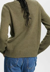 Pull en maille vert olive avec un motif en nid d'abeille texturé, un col, des poignets et un ourlet côtelés ; associé à un jean en denim bleu.