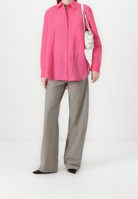 Helles rosa Hemd mit Knöpfen und einer zerknitterten Textur sowie Tasche, kombiniert mit hellgrauen, weiten Hosen und schwarzen spitzen Absatzschuhen.