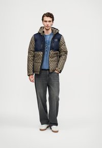 Jovem vestindo uma jaqueta com padrão castanho e azul marinho com capuz, uma t-shirt gráfica azul, uns jeans soltos cinzentos escuros, e ténis brancos, em pé.