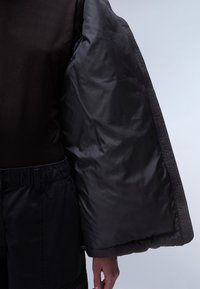 Czarna pikowana kurtka z połyskującą, pikowaną fakturą. Rękaw jest szeroki i bufiasty, zestawiony z dopasowanym ciemnym topem i czarnymi spodniami.