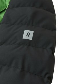 Giacca a vento nera in tessuto con finitura testurizzata, dotata di un logo grigio su una toppa quadrata. Il design include cuciture visibili e un rivestimento verde.