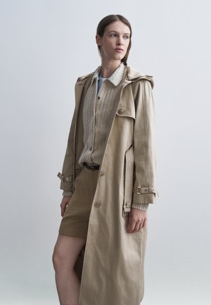 Frau steht und trägt einen beigen Trenchcoat über einer gerippten Strickjacke, einem Hemd mit verzierter Kragen und passenden beigen Shorts vor einem einfarbigen Hintergrund.