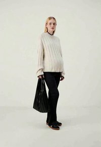 Strickpullover in Übergröße in Creme mit gerippter Textur, schwarze Kante, kombiniert mit schwarzen Hosen und einer gefalteten schwarzen Tasche. Das Model steht vor einem hellen Hintergrund.