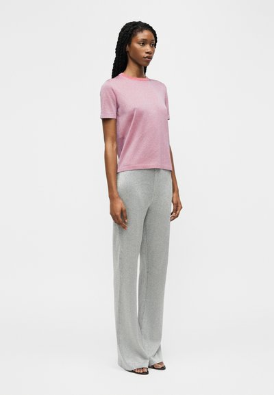 Γυναίκα με μακριά πλεξούδα φοράει ροζ μπλουζάκι με κοντά μανίκια και γκρι παντελόνα ψηλόμεση με φαρδύ πόδι, στέκεται μπροστά σε απλό φόντο.