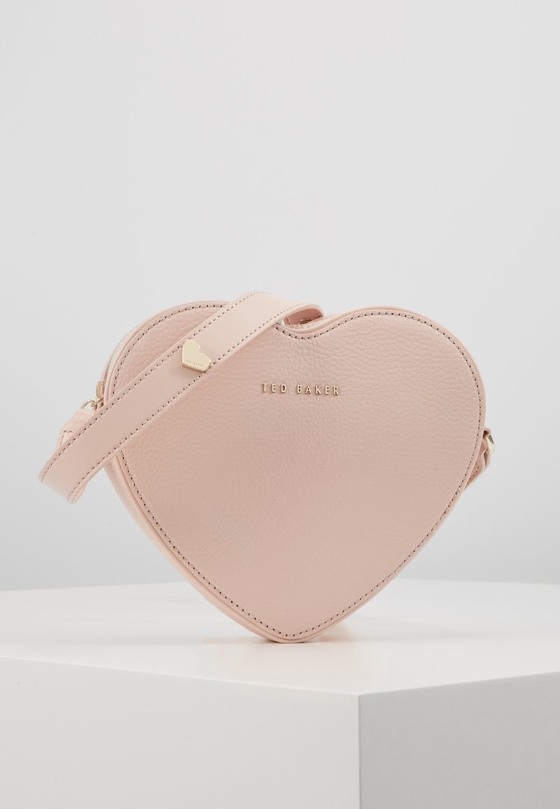 Torebka w kształcie serca w różowym kolorze, wykonana z gładkiej skóry, z złotym logo, gładką fakturą oraz odczepianym paskiem z małym złotym akcentem w kształcie serca.