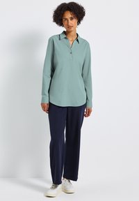 Camicia a maniche lunghe verde chiaro con colletto, patta di bottoni e vestibilità rilassata, abbinata a pantaloni a gamba larga blu navy e sneakers bianche.