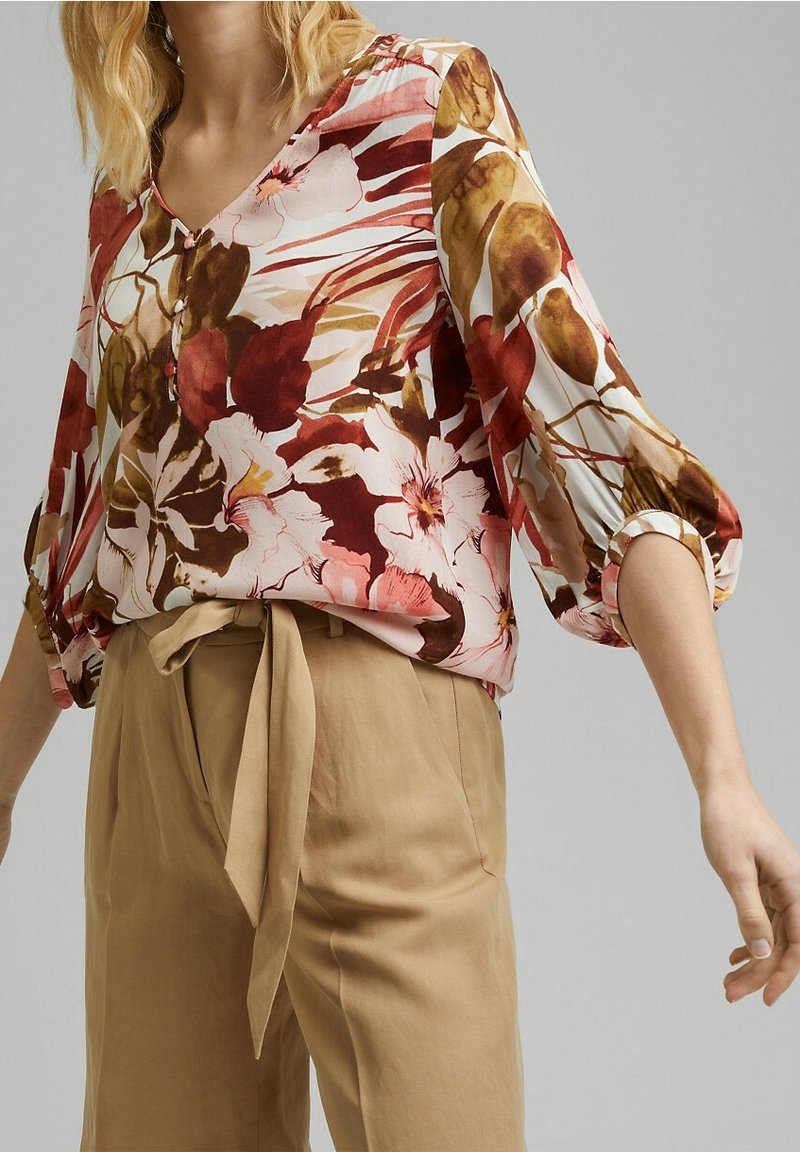 Blusa floreale bianca, rosa e marrone con scollatura a V, maniche a tre quarti e dettagli con bottoni. Abbinata a pantaloni beige a vita alta.