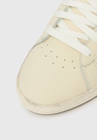 Κοντινό πλάνο της μύτης ενός μπεζ αθλητικού παπουτσιού με λευκά κορδόνια, μικρές τρύπες διάτρησης και ελαφρώς φθαρμένη υφή στη σόλα.