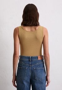 Vrouw met schouderlang bruin haar, gekleed in een mouwloze beige top en hoog waaiers blauwe jeans, staand tegen een eenvoudige witte muur.
