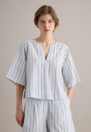 Woman wearing loose white top and matching wide-leg pants with vertical blue stripes, standing against a plain light background.