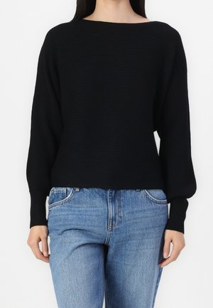 Vrouw draagt een zwart geribd trui met lange mouwen en lichtblauwe hoge taille jeans, staand tegen een effen witte achtergrond.