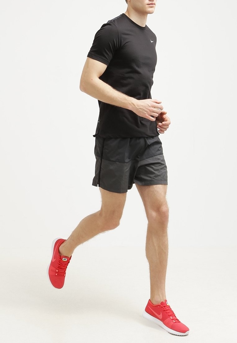 Homme en jogging portant un t-shirt noir, un short gris foncé et des chaussures de course Nike rouge vif sur un fond blanc uni.